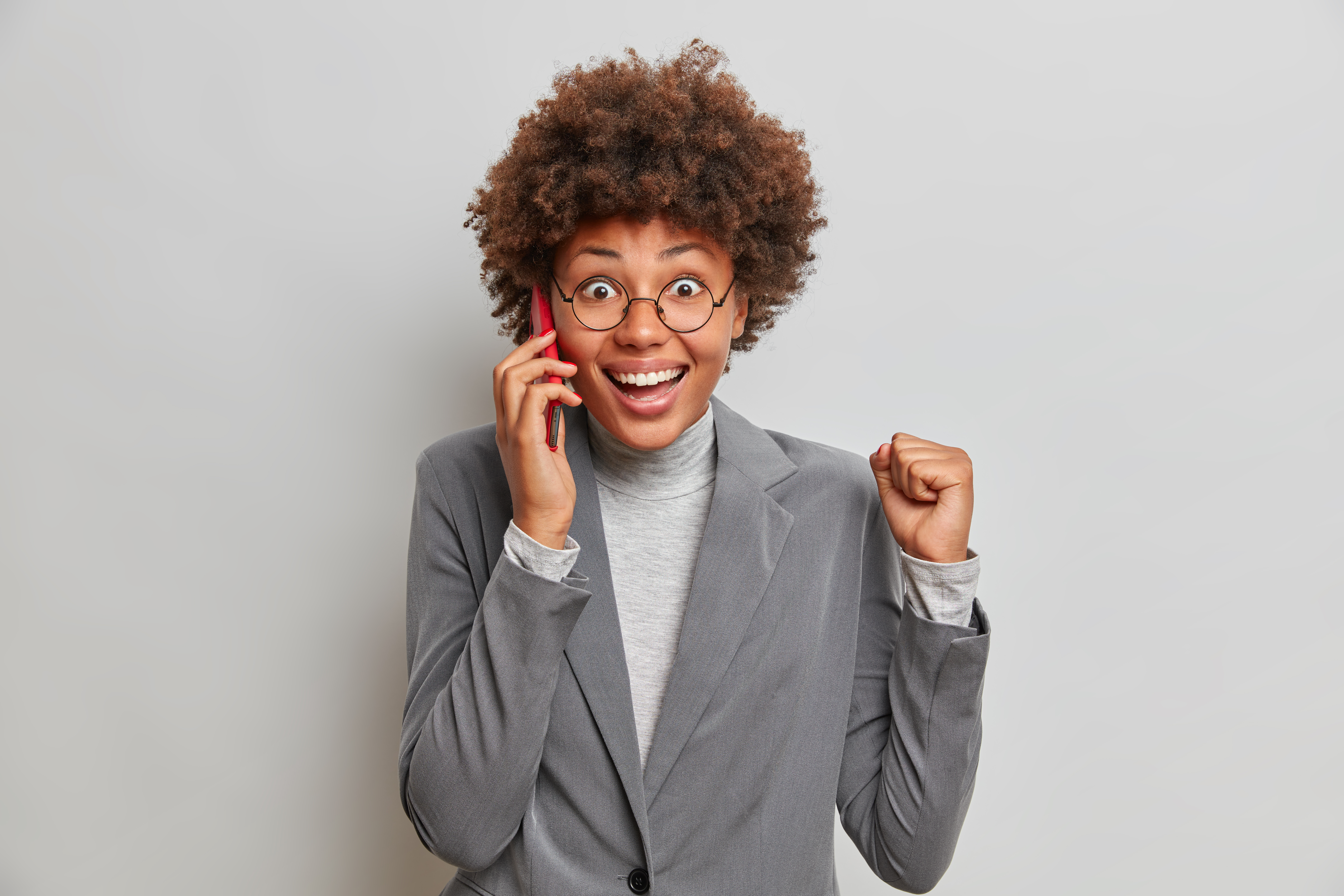 Overjoyed successful business lady with Afro hair celebrates successful deal, raises clenched fist as talks with partner via mobile phone. Joyful female manager receives positive feedback grom client